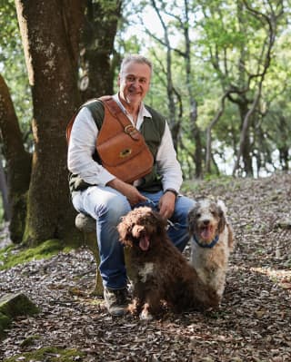 A smiling man sits on a rock in the woods, a leather saddle back slung across a shoulder. He hold the leads of two happy dogs