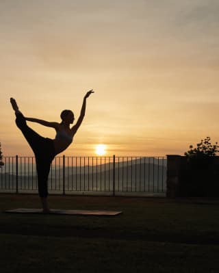 A young woman stands on a yoga mat in a perfectly balanced dancer pose as the sun slowly climbs into dawn behind her