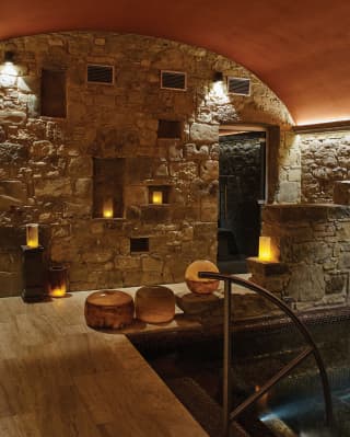 Large candles fill the alcoves of a stone wall below an arched terracotta ceiling, illuminating a deep mosaic pool