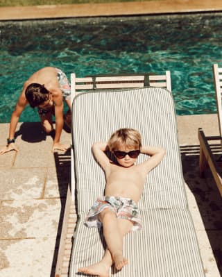 Frecher Junge mit Badehose und Sonnenbrille, der sich auf einer Sonnenliege zurücklehnt 