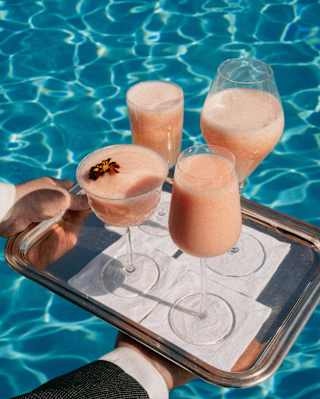 Close-up on a tray of Peach Bellini cocktails served in four different-shaped glasses with a shimmering pool-water backdrop.