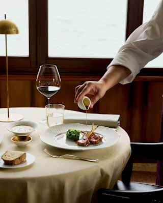 A waiter pours unctuous gravy from a small white jug onto a dish of delta lamb with chicory, in a view of a table at Oro.