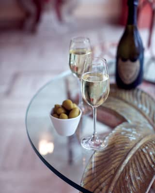 A bottle of Prosecco stands on a glass table beside two filled champagne flutes with a small ceramic bowl of green olives