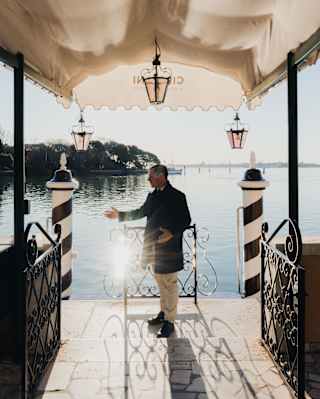 Sun dazzles on the water in front of the hotel, where a staff member in a black jacket stands between the monochrome paline.