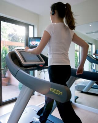 A woman in a white t-shirt and black yoga pants is reflected in mirrors as she strides on a treadmill in a well-equipped gym