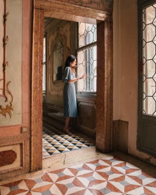 A woman in a soft pleated skirt looks through tall leaded windows in an ancient Venetian palace with brightly tiled floors