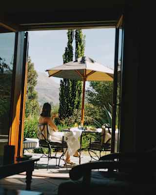 A woman sits at a terrace table with coffee, shaded by a parasol, facing the views, seen from a room interior through a door.