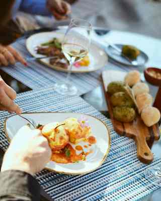 Two guests enjoy al-fresco lunch at a Curiña table with vibrant dishes, a plank of breads and white wine on blue raffia mats.