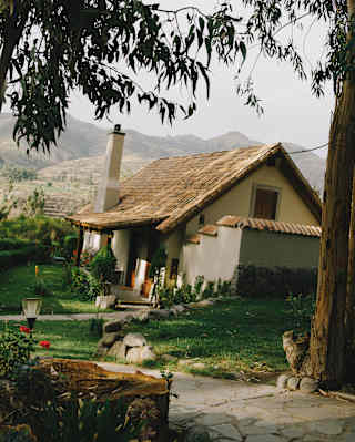 Slightly angled shot of a residence with a steep-gabled roof, tucked into the Las Casitas gardens, framed by tall trees.