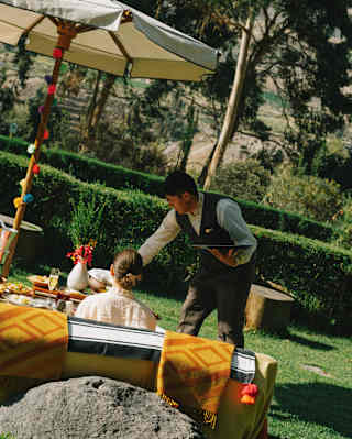 Angled image of a waiter holding a tray in one hand and serving a drink with the other to a woman enjoying a garden lunch.