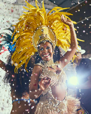 Lady in a glittering gold carnival outfit with a grand yelllow-feathered headpiece dancing among confetti 