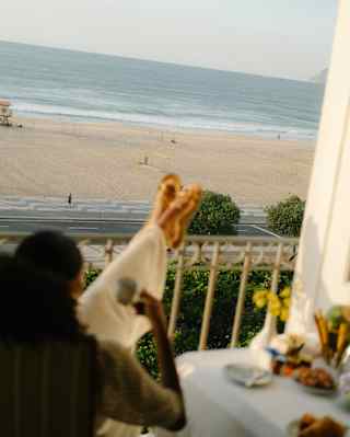 In soft-focus foreground a woman drinks coffee with her feet up on balcony railings, gazing at the calm beach below.