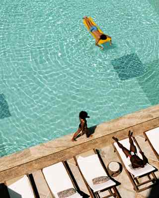 Aerial view of a woman walking the edge of the pool, where a man on his front on a yellow lilo creates light-dappled ripples.