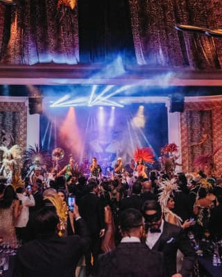 A ballroom is in the mood to party, with samba dancers in feathered crowns, a live band and revellers in carnival masks