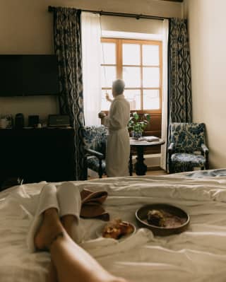 A guest stretches her legs on a bed alongside plates of pastries, as a man in a robe adjusts drapes to let in morning light.