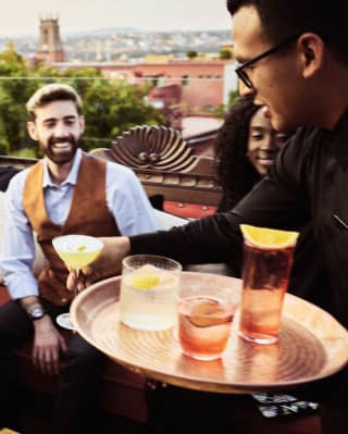 A smiling group are served cocktails on sofas at the Tunki bar with rooftop views of the historic city behind