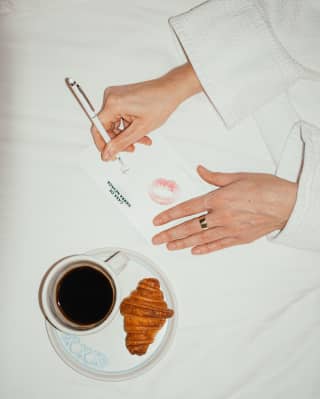 Birdseye view of a female guest penning a lipstick-marked note on hotel-headed paper at a table with croissant and coffee.