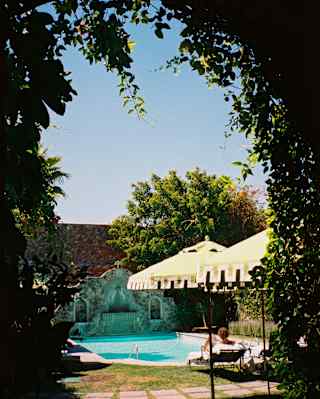 A guest relaxes on a chair by the sparkling pool in the hotel's walled garden, seen through an arch clad with climbers.