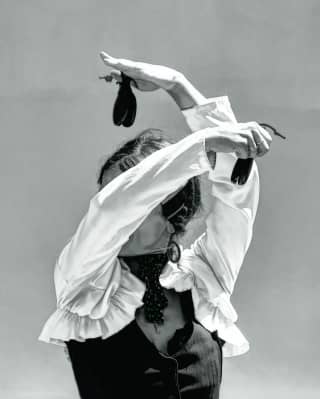A female flamenco dancer raises her arms in front of her face, holding castanets - shot in black and white from the waist up.