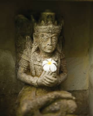 Close-up of a carved stone Hindu deity in an alcove shrine, holding a freshly-placed offering of a frangipani flower.