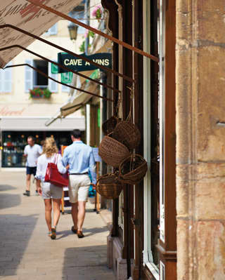 View along shop fronts in a French town, with baskets tied to awning supports and signs saying Fromagerie and Cave a Vins.