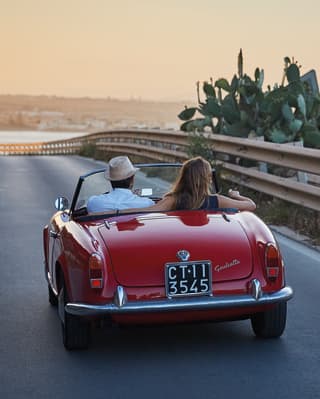 Gleaming, red, soft-top classic car on a seaside road at sunset