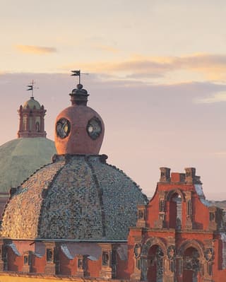 Sundown on the roofscape of central San Miguel de Allende, with mosaic domes in green and grey, and turrets in red and pink.