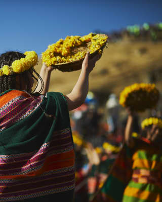 A woman in a woven shawl with long braids and yellow marigold crown, holds a basket of flowers, celebrating the Sun Festival.