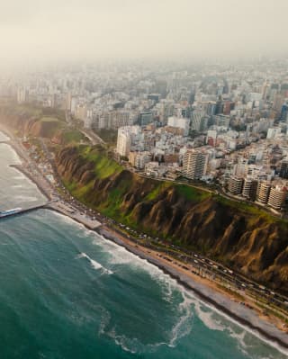 Ariel view of Lima atop dramatic green cliffs that plunge down to the Pacific coastline, where a pier reaches into the ocean.