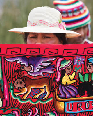 A local woman holds up a handwoven red rug, embroidered with depictions of Peruvian culture and folklore in bright threads.