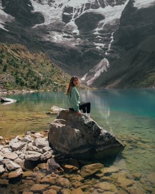 A young woman in poses on rock by the sapphire lagoon of Humantay Lake, cupped between snowy mountains in the highlands.