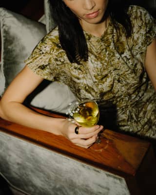 A woman with dark hair in a green print reclines on a sofa holding a Gin and Jazz Negroni Blanco cocktail, seen from above.