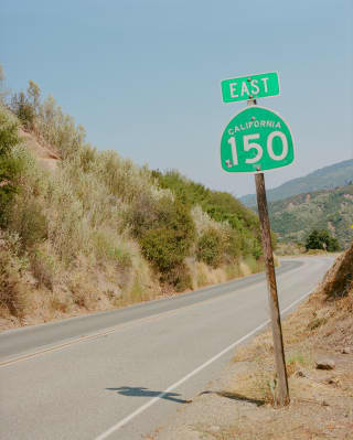 A signpost saying East and California 150 in green paint tilts slightly in the dusty roadside casting a shadow on the road.