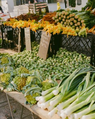 An market stall sells leaks, cabbages and Brussel sprouts, with a shelf above overflowing with produce and courgette flowers.