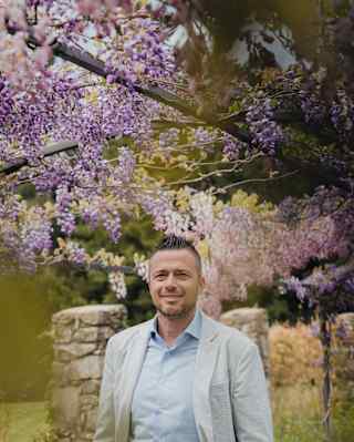 Wearing a light suit, Alessandro Baccarelli, General Manager at Castello Di Casole, stands in gardens under a lilac wisteria.