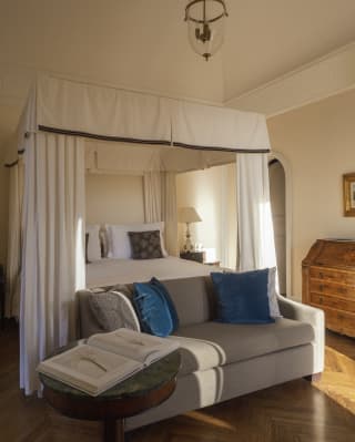 A beige sofa sits at the end of a four-poster bed with white canopy which offers a palatial feel to the Floresta Suite.