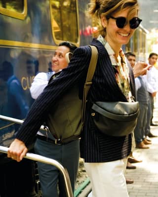 Train staff line the platform as a woman with blond hair, wearing sunglasses, black blazer and white trousers, disembarks.