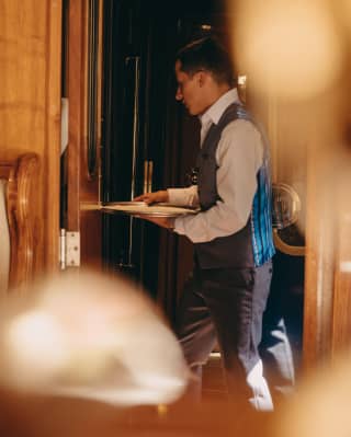 A partly obscured image of a waiter, wearing and blue and black waistcoat, as he carries a round silver tray through a carriage door.