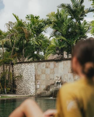 In soft-focus foreground, a woman in a yellow gown relaxes, gazing at the pool in the walled garden overlooked by tall palms.