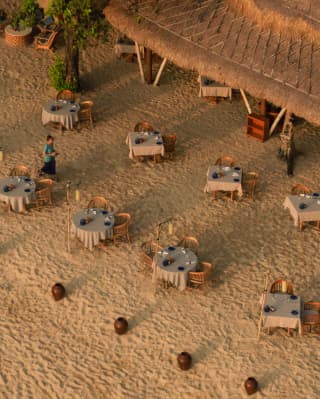 A waitress walks between ten tables placed on the rippled sand outside Neyalan, ready for sunset dining, seen from above.