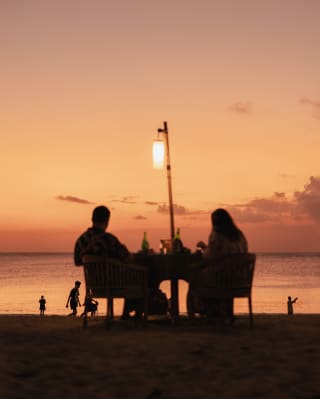 A silhouetted couple sits at a beachside table with a lantern, watching children play by the sea against a peach sunset sky.