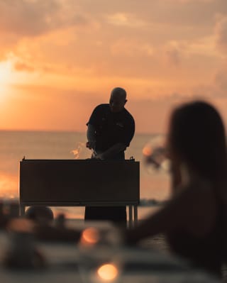 Silhouetted against the peachy sunset, a chef prepares food on a beach barbeque watched by a woman in soft-focus foreground.