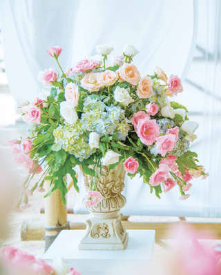 A floral bouquet of pink, cream and white roses with bursts of greenery, sprays from an ornately-carved flower urn.