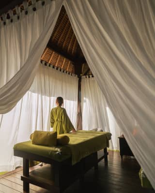 A woman in an olive-green gown stands by a treatment bed at the Beach Spa, seen from behind, through open white drapes.