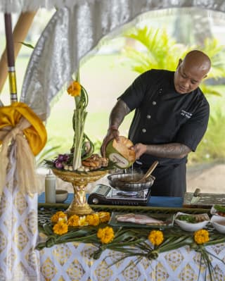 Chef Adi demonstrates the cooking of Young Coconut Water Seafood Soup at a table loaded with produce, with floral decoration.