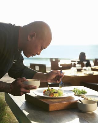 Executive Chef Adi Wijaya, in black uniform, carefully spoons dressing over a vibrant, fresh dish at a beachside table.