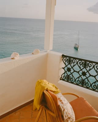 An empty chair sits in a balcony corner where wall meets rail, offering extensive views of the sea and a single sailing boat.