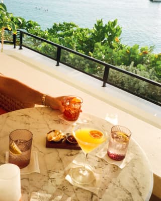 At Baie Longue Bar, a guest's hand reaches for a tumbler at a table with aperitifs and cocktails, with treetop and sea views.