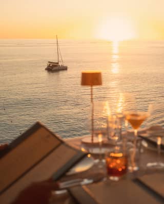 In soft-focus, an off-camera guest peruses the L'Oursin menu at an outside table, as the sun sets on the ocean horizon.