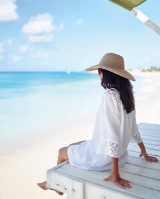 A woman in a white cotton tunic and floppy straw hat dangles her feet over the wooden deck of her beachside cabana